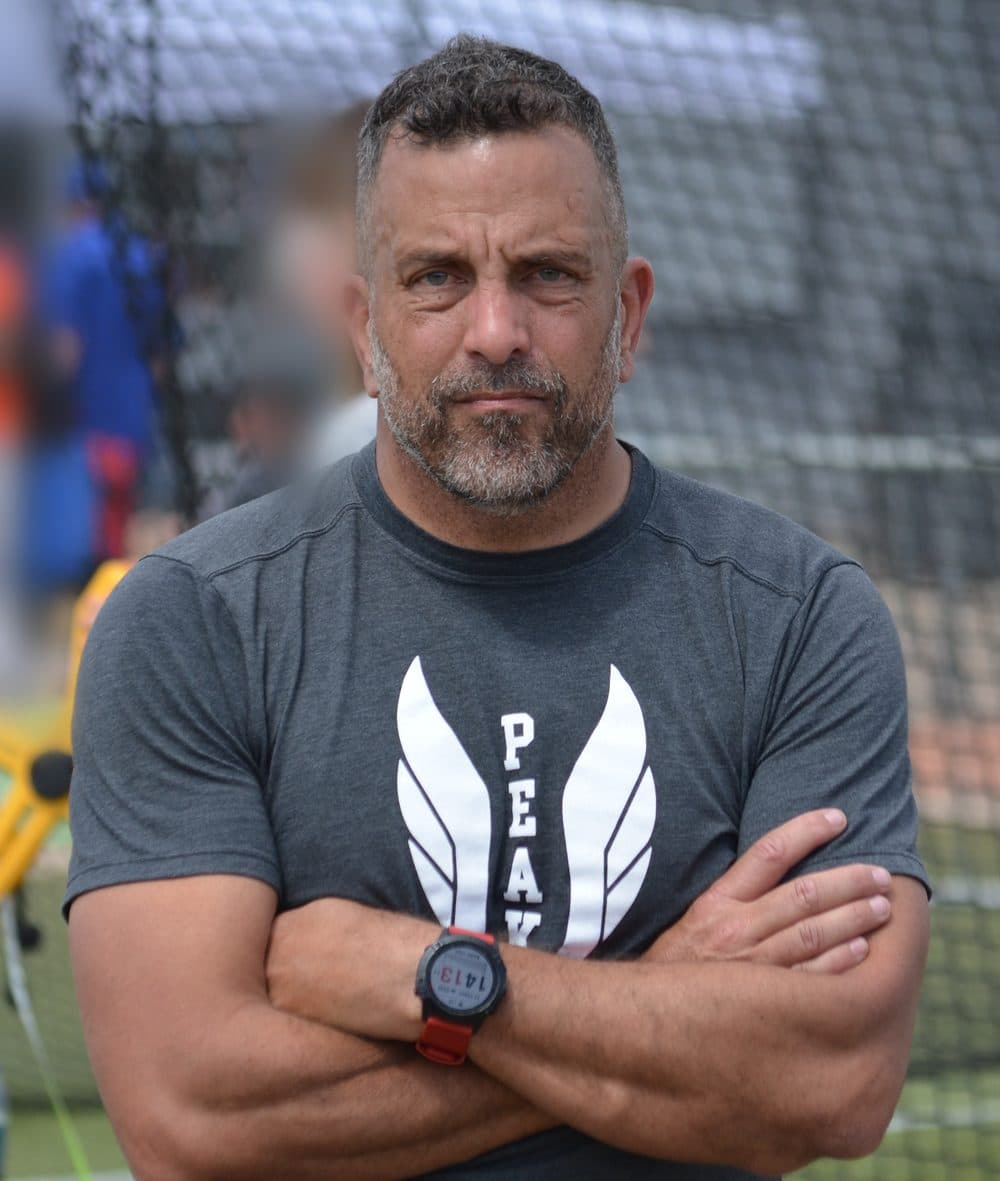 Brandon Watson, founder of OfferSecured, standing with arms crossed beside a throwing cage at a high school track meet, wearing a Peak to Peak track & field shirt.